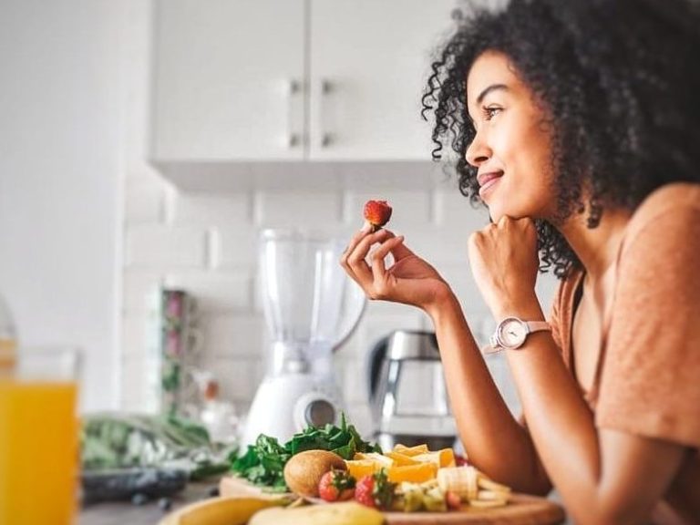 "Young woman leaning on the kitchen counter, enjoying a strawberry, and surrounded by fruit, looking thoughtful and smiling."