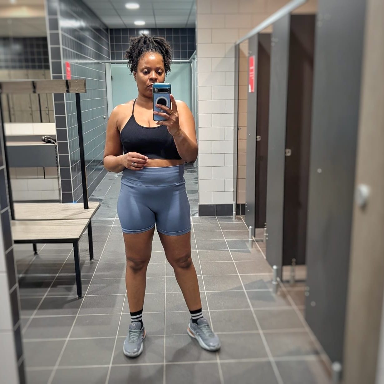 "Woman in gym changing room wearing high-waisted shorts and a black crop top showing the real starting point of her fitness journey." 