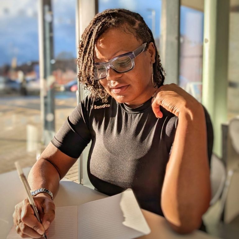 "Woman writing thoughtfully in a diary in a bright room, wearing blue glasses, looking down as she reflects on her wellness journey."