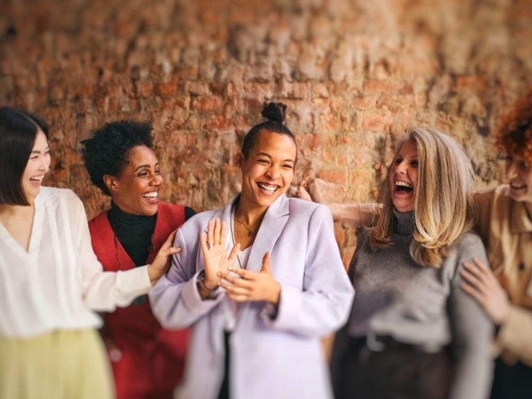 "A diverse group of six women sitting outdoors in the sunshine, smiling and talking together."