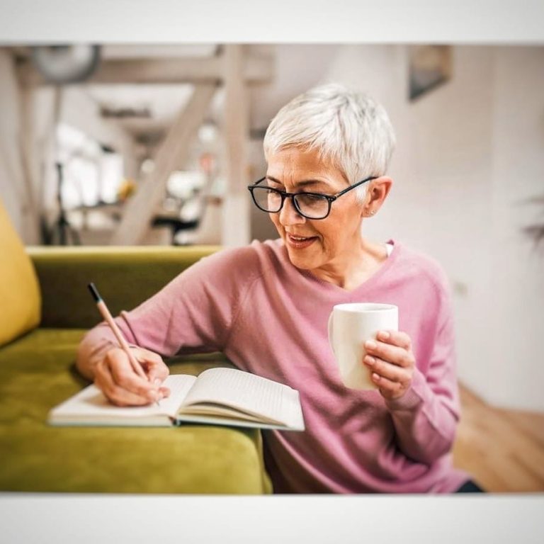 "Women sitting indoors and writing in a notebook with a cup in her hand, appearing thoughtful and focused."